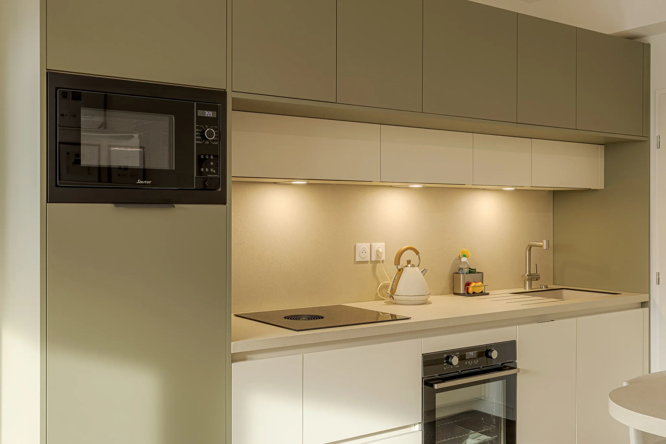 Cuisine compacte sur mesure avec colonne intégrant le micro-ondes, rangements hauts vert sauge et plan de travail clair
