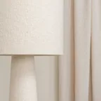 Détail d’un abat-jour en tissu beige devant des rideaux clairs, pour une ambiance douce et chaleureuse.