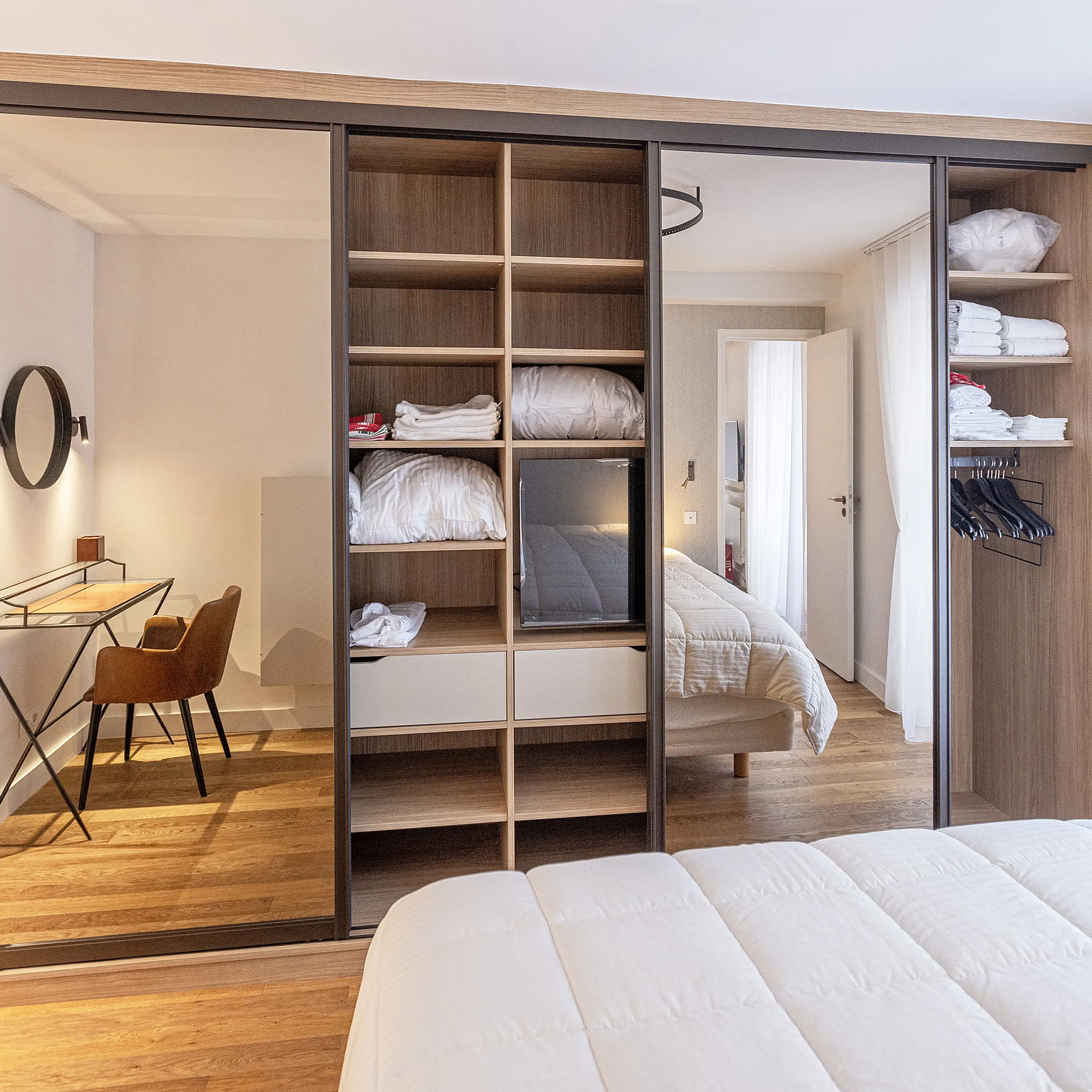 Aménagement de chambre avec dressing sur mesure, niches ouvertes, portes miroir et espace bureau intégré sur le côté.