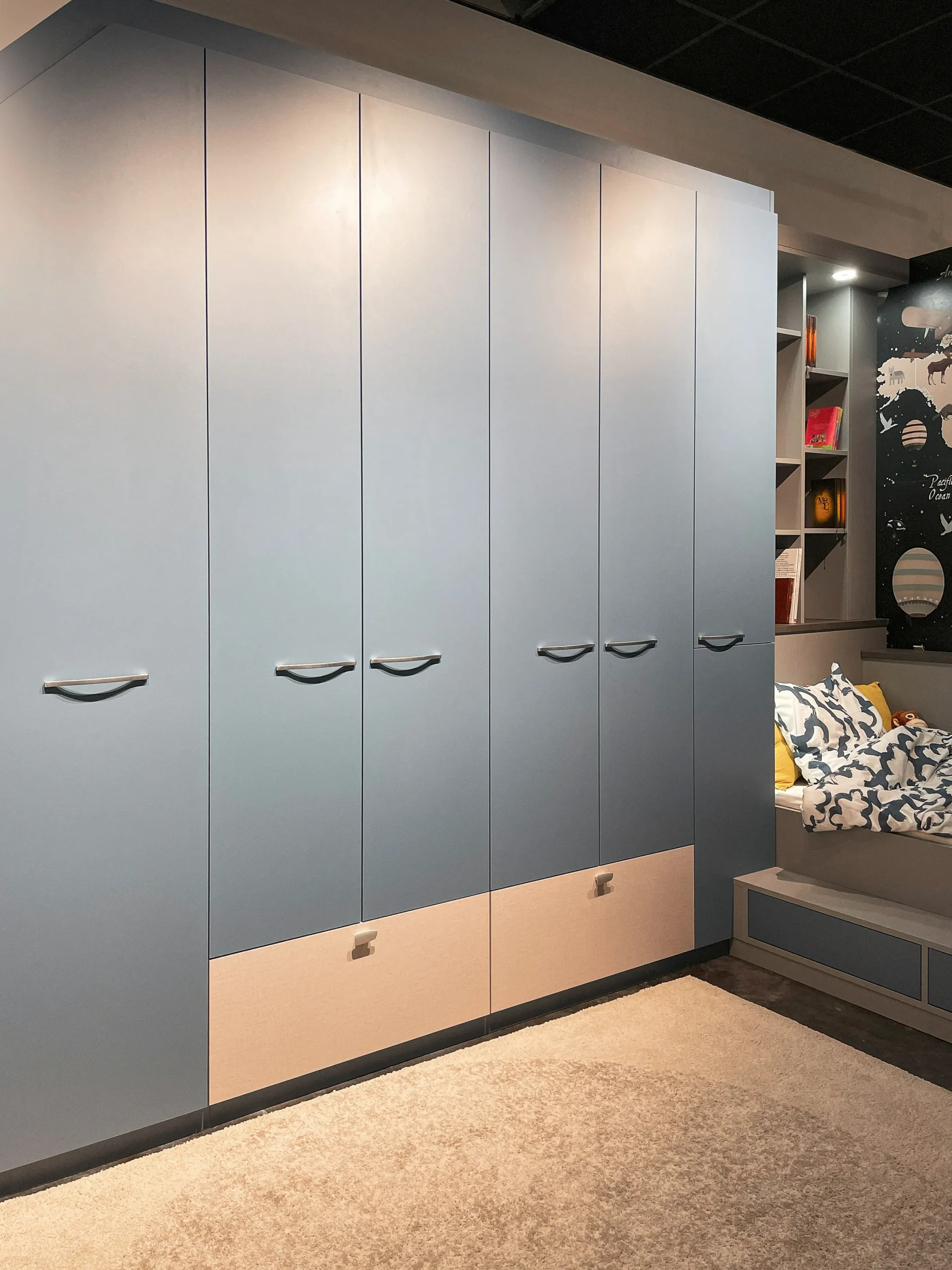 Armoire murale bleue sur-mesure avec tiroirs clairs, placards hauts et lit enfant intégré près d’un décor carte du monde.