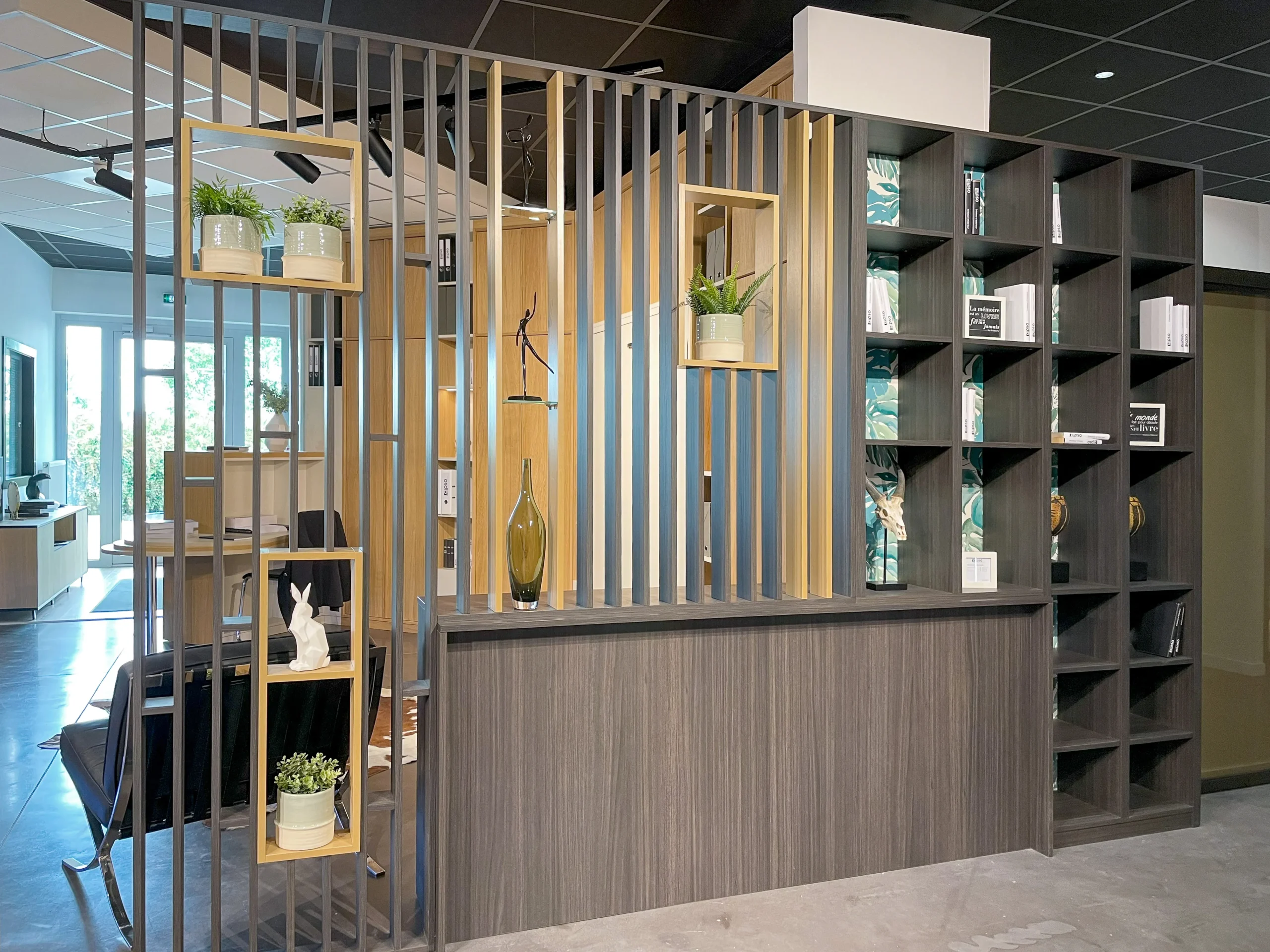 Grand claustra bibliothèque en bois foncé et bois clair, avec niches végétalisées, étagères ouvertes et séparation ajourée entre deux espaces.