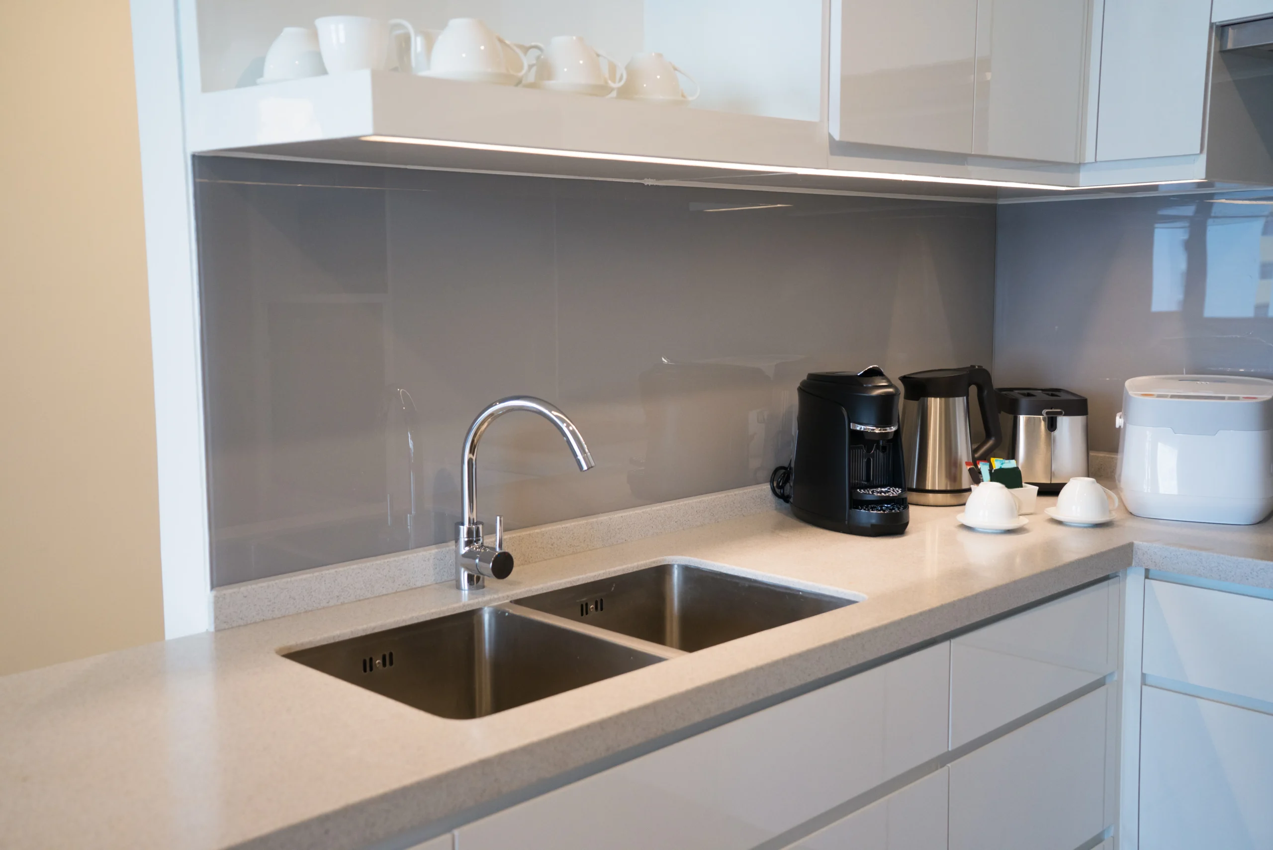 Cuisine blanche contemporaine avec évier double inox, crédence grise brillante, plan de travail clair et petit électroménager intégré.