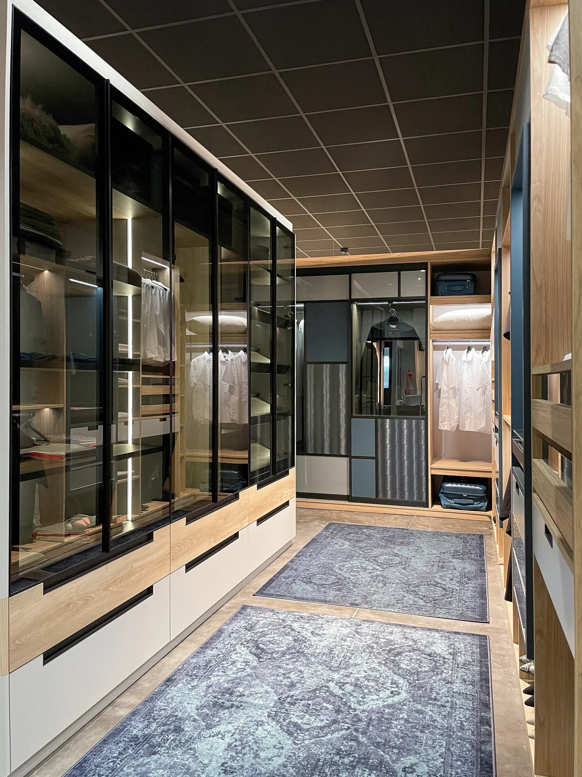Dressing sur-mesure en bois clair avec façades vitrées noires, penderies éclairées, rangements bas et modules ouverts pour organiser vêtements et chaussures.