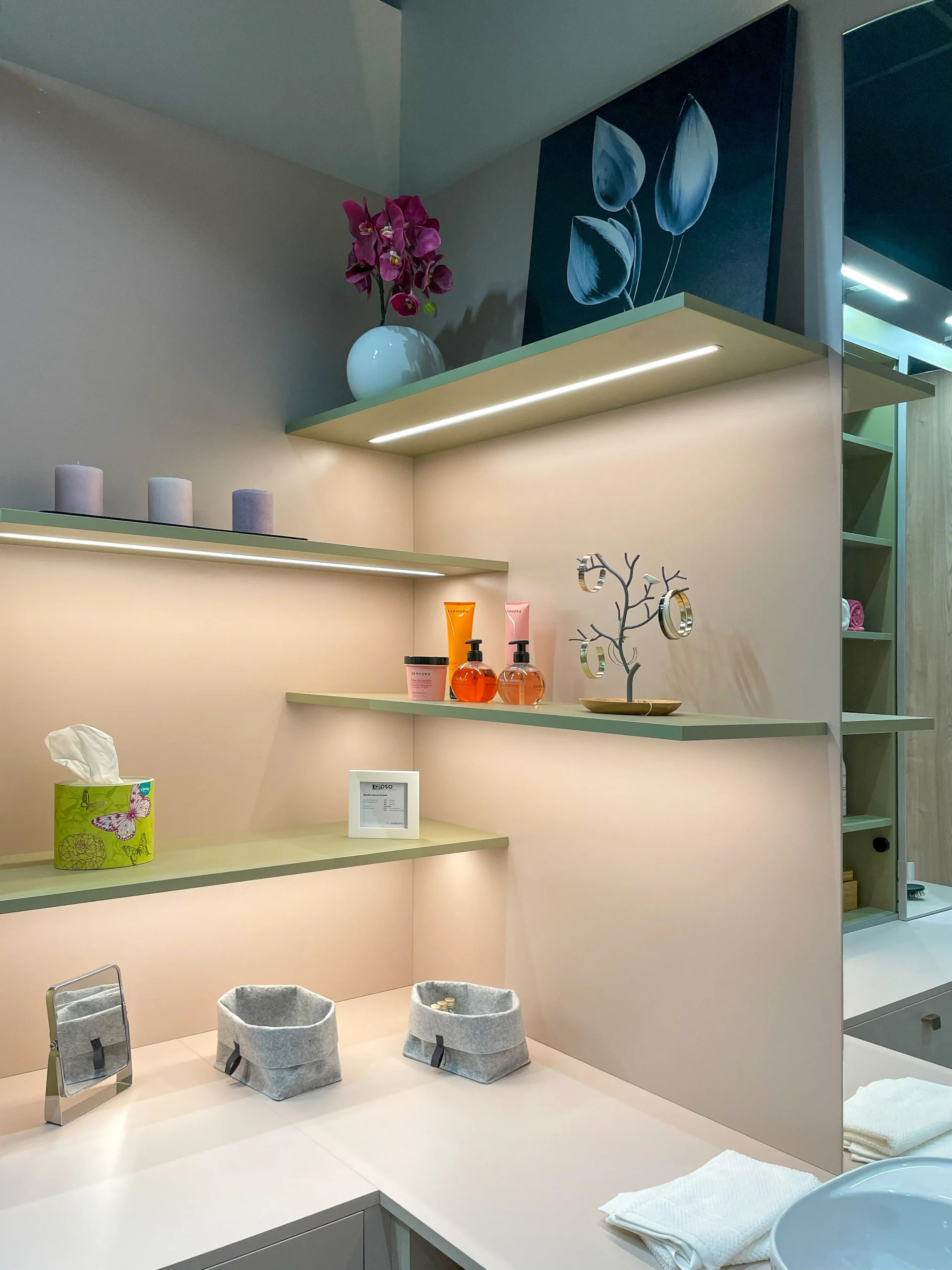 Détail d’étagères murales éclairées avec accessoires de toilette, finitions vert sauge et fond rose poudré.