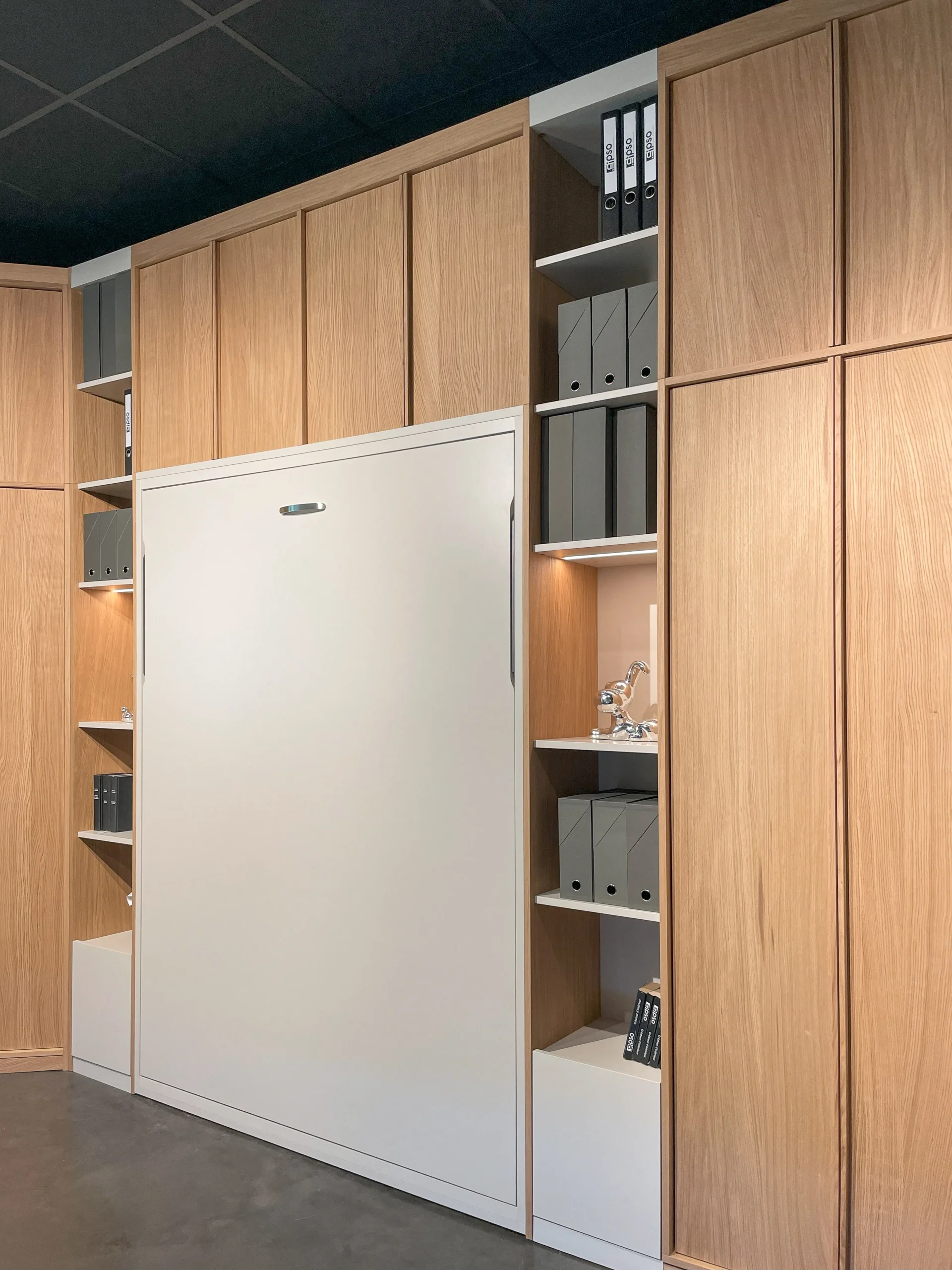Vue rapprochée d’un lit escamotable fermé, encadré par des rangements muraux en bois clair et des niches décoratives.