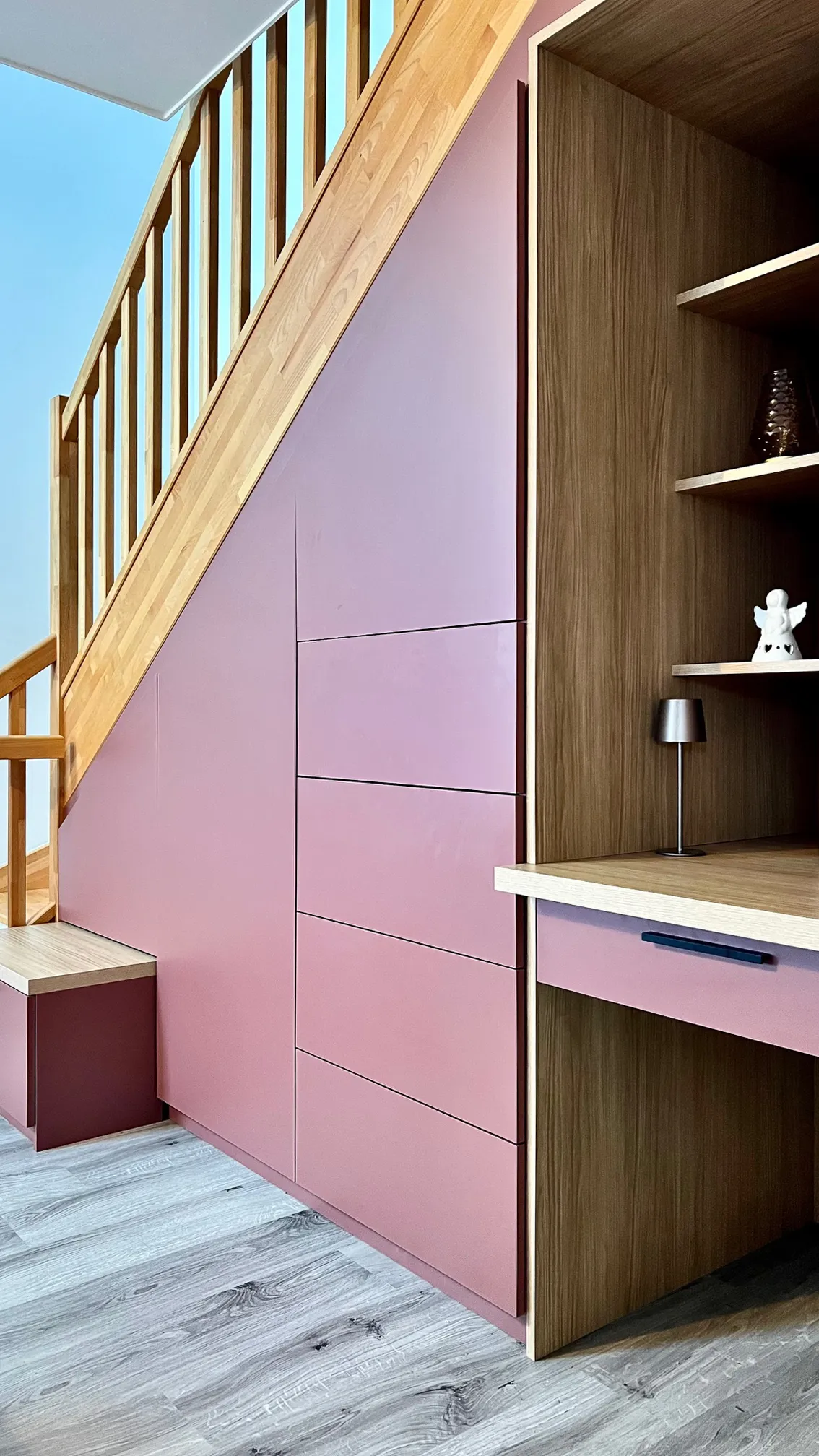 Aménagement sous escalier rose et bois avec placards, tiroirs, niches ouvertes et bureau intégré.
