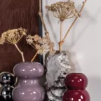 Composition de vases décoratifs rose, bordeaux et argent avec fleurs séchées, posés sur un meuble dans une ambiance chaleureuse.