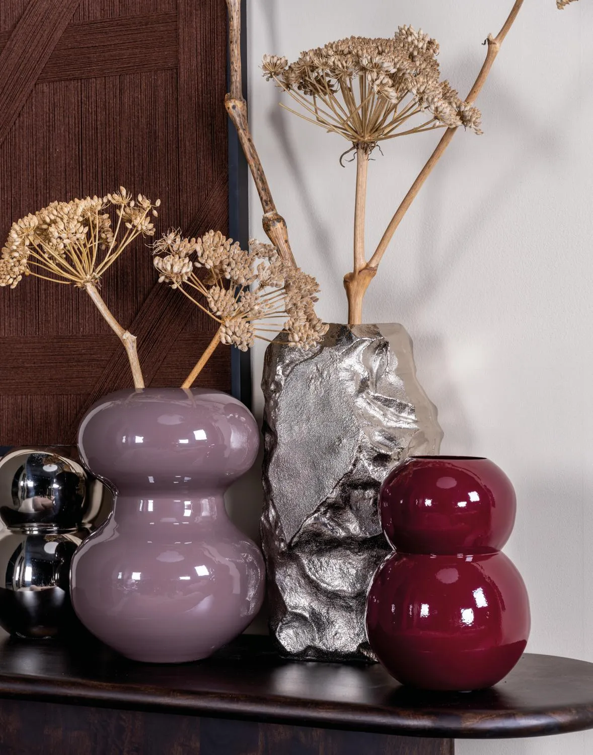 Composition de vases décoratifs rose, bordeaux et argent avec fleurs séchées, posés sur un meuble dans une ambiance chaleureuse.